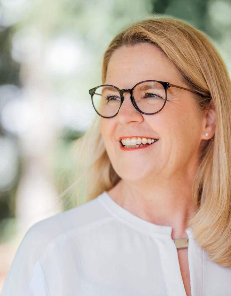 Meike Schuettkus Freie Trauerrednerin freundlich laechelnd An Ihrer Seite: Meike Schüttkus Freie Trauerrednerin, hier in weißer Bluse, blonde Haare, Braune Brille
